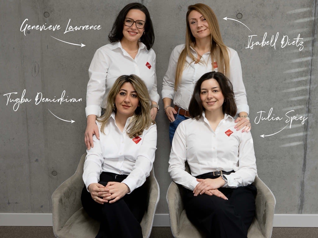 Gruppenfoto eines vierköpfigen Recruiting‑Teams vor einer grauen Wand. Zwei Personen stehen in der hinteren Reihe, zwei Personen sitzen davor auf Sesseln. Alle tragen weiße Oberteile und dunkle Hosen. Die Personen stehen bzw. sitzen nah beieinander und wirken als Team arrangiert.