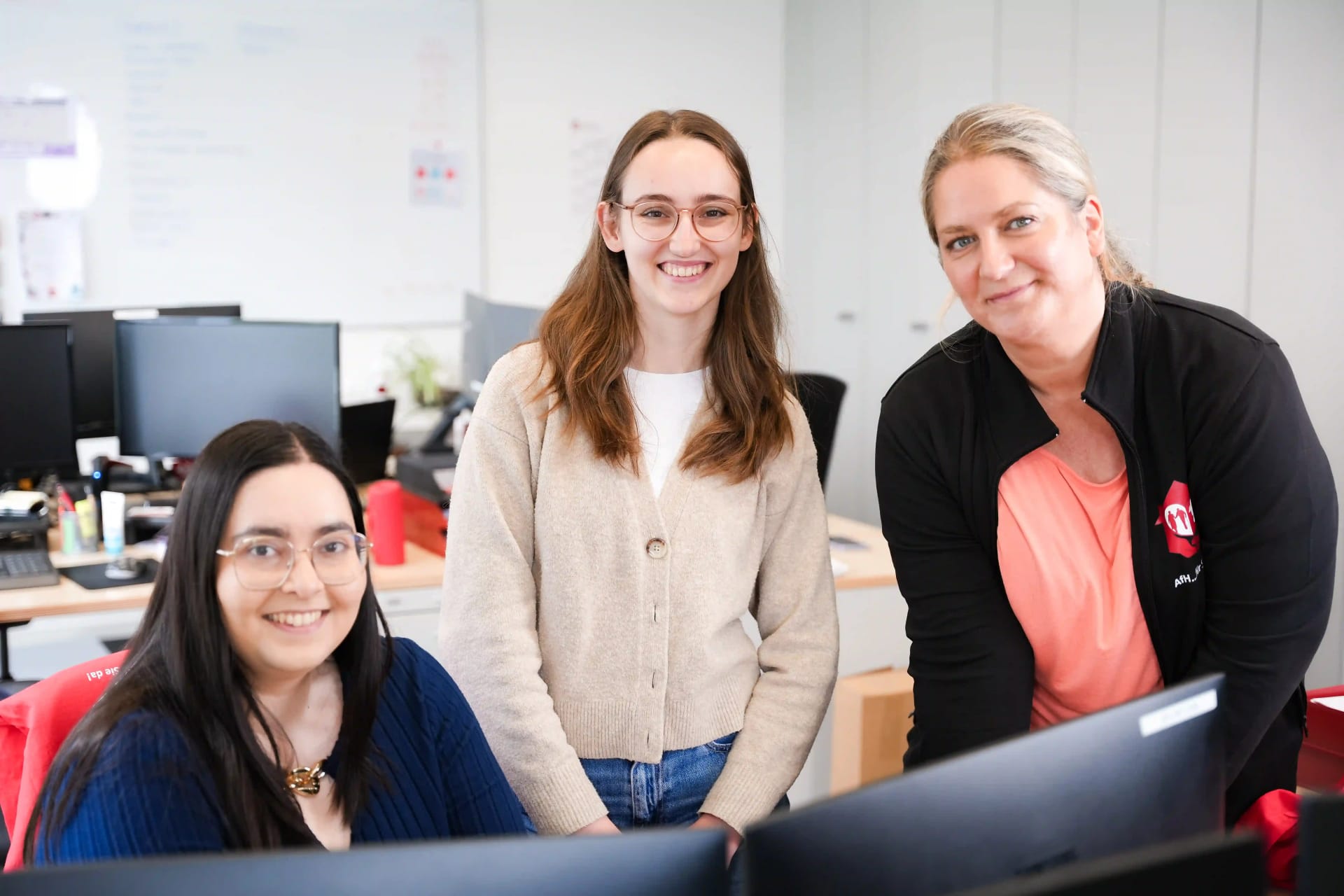 Drie medewerksters van het marketingteam van het Agency For Domestic Help staan en zitten samen glimlachend aan hun bureau.