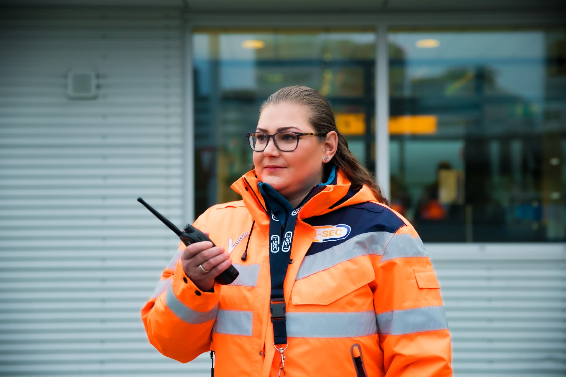 Vrouwelijke beveiligingsmedewerker met een oranje hi-vis jas communiceert via een walkietalkie buiten een kantoorgebouw.