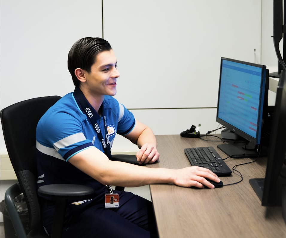 Jonge mannelijke I-SEC beveiligingsmedewerker met badge werkt aan een bureau met een computer in een kantooromgeving.