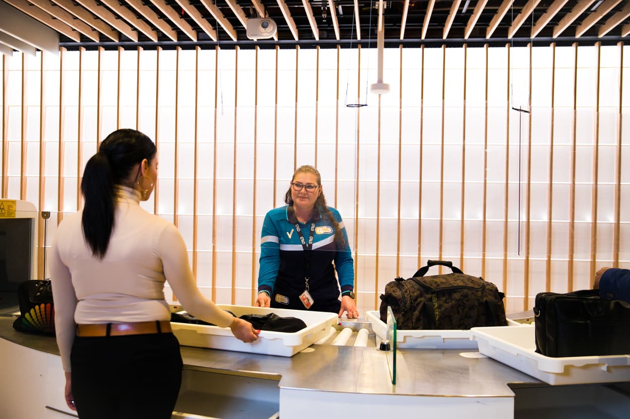 Vrouwelijke I-SEC beveiligingsmedewerker controleert de inhoud van een bakje bij een luchthavenveiligheidscontrole terwijl een passagier wacht.
