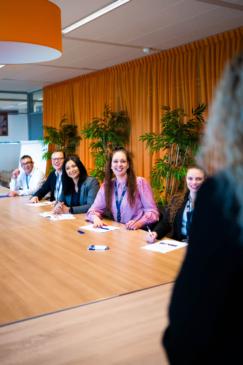 Drie vrouwelijke collega's lachen en zitten aan een vergadertafel in een helder kantoor.