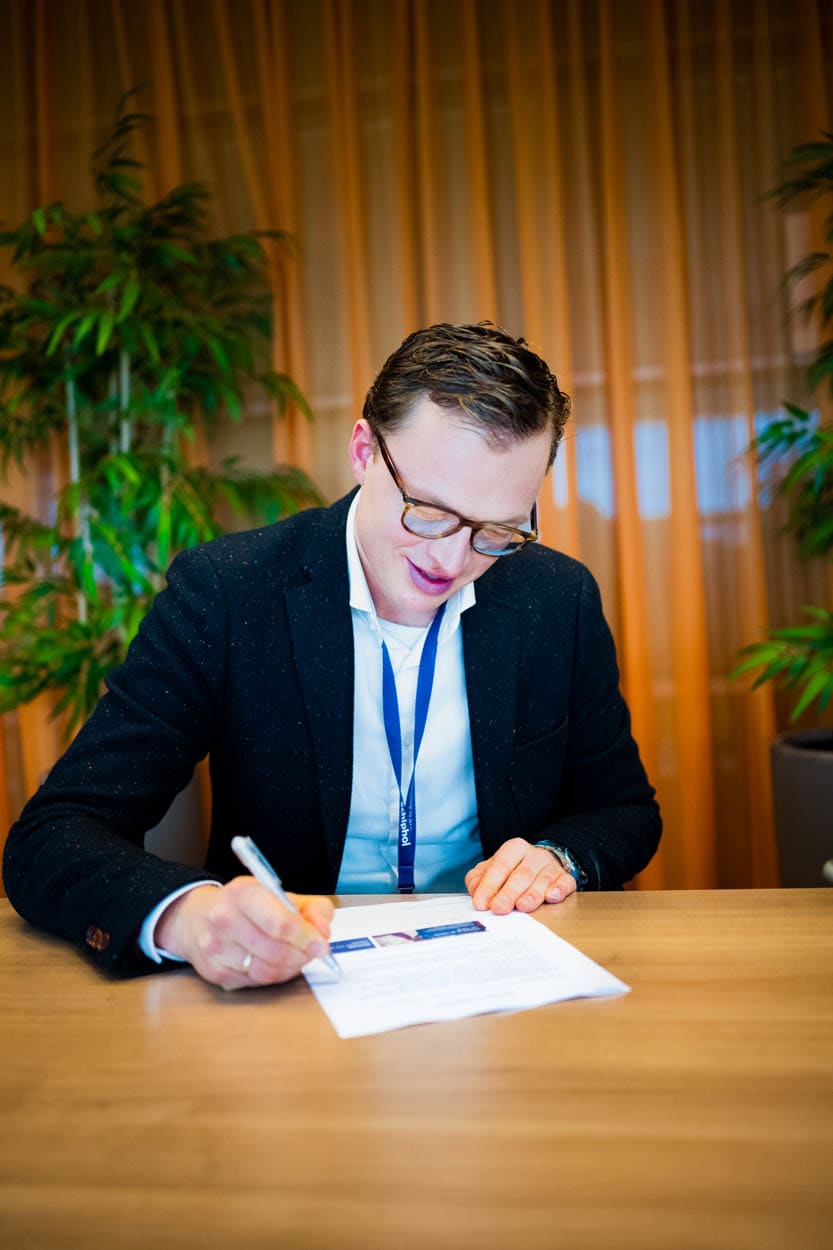 Een jonge mannelijke professional schrijft aantekeningen, zittend aan een kantoor werkplek met een laptop.
