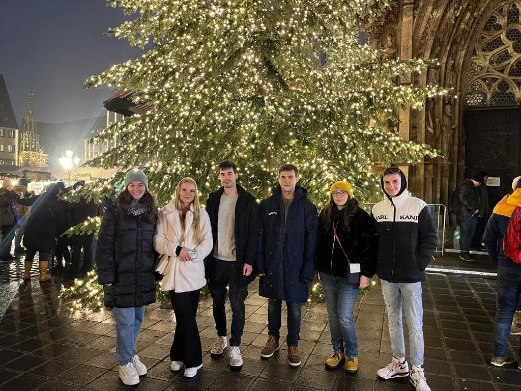 Teamfoto der Auszubildenden der Firma Walraven auf dem Bayreuther Weihnachtsmarkt vor einem großen, beleuchteten Weihnachtsbaum