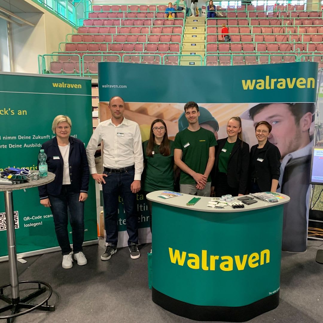 Drei Azubis sowie drei weitere Mitarbeiter der Walraven GmbH auf der Ausbildungsmesse in der Oberfrankenhalle in Bayreuth. Alles sechs Personen stehen vor einer Rückwand, neben zwei grünen RollUps und gleichzeitig hinter einer grünen Theke mit Walraven-Logo. Im Hintergrund sind die Tribünen der Halle zu sehen.