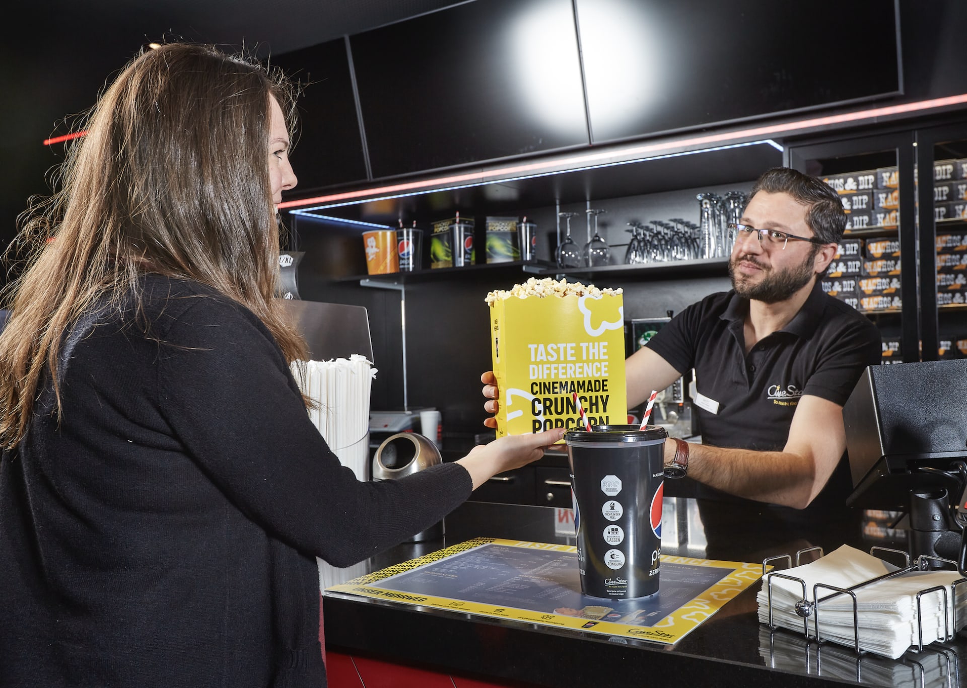 Ein CineStar Mitarbeiter übergibt einer Gästin Popcorn, Die Szene spielt an einer Kinokasse mit Snacktheke. 