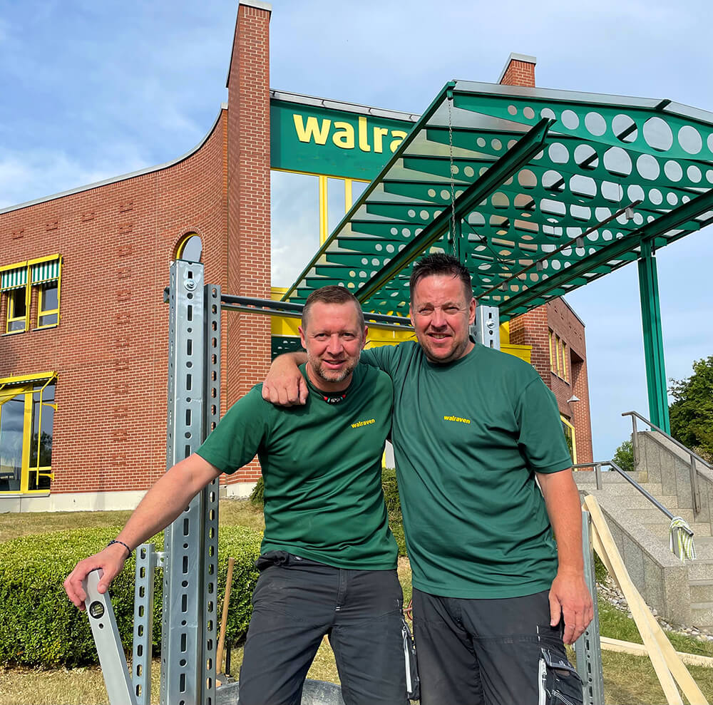 Zwei Mitarbeiter der Walraven GmbH in Bayreuth stehen vor dem Haupteingang, lachen in die Kamera und haben sich gegenseitig im Arm.