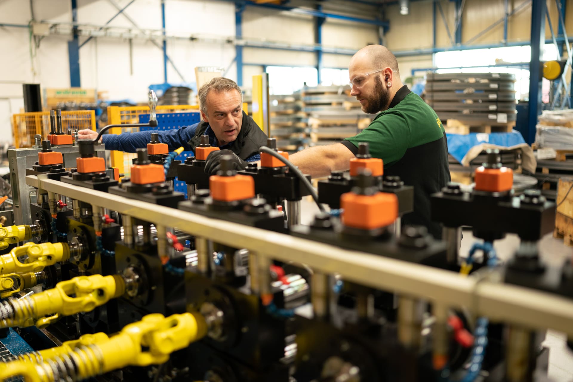 Abbildung zweier Walraven-Mitarbeiter in der Produktionshalle in Mijdrecht. Sie sprechen miteinander und prüfen bei der entsprechenden Maschine, ob die Herstellung der Montageschienen reibungslos abläuft.
