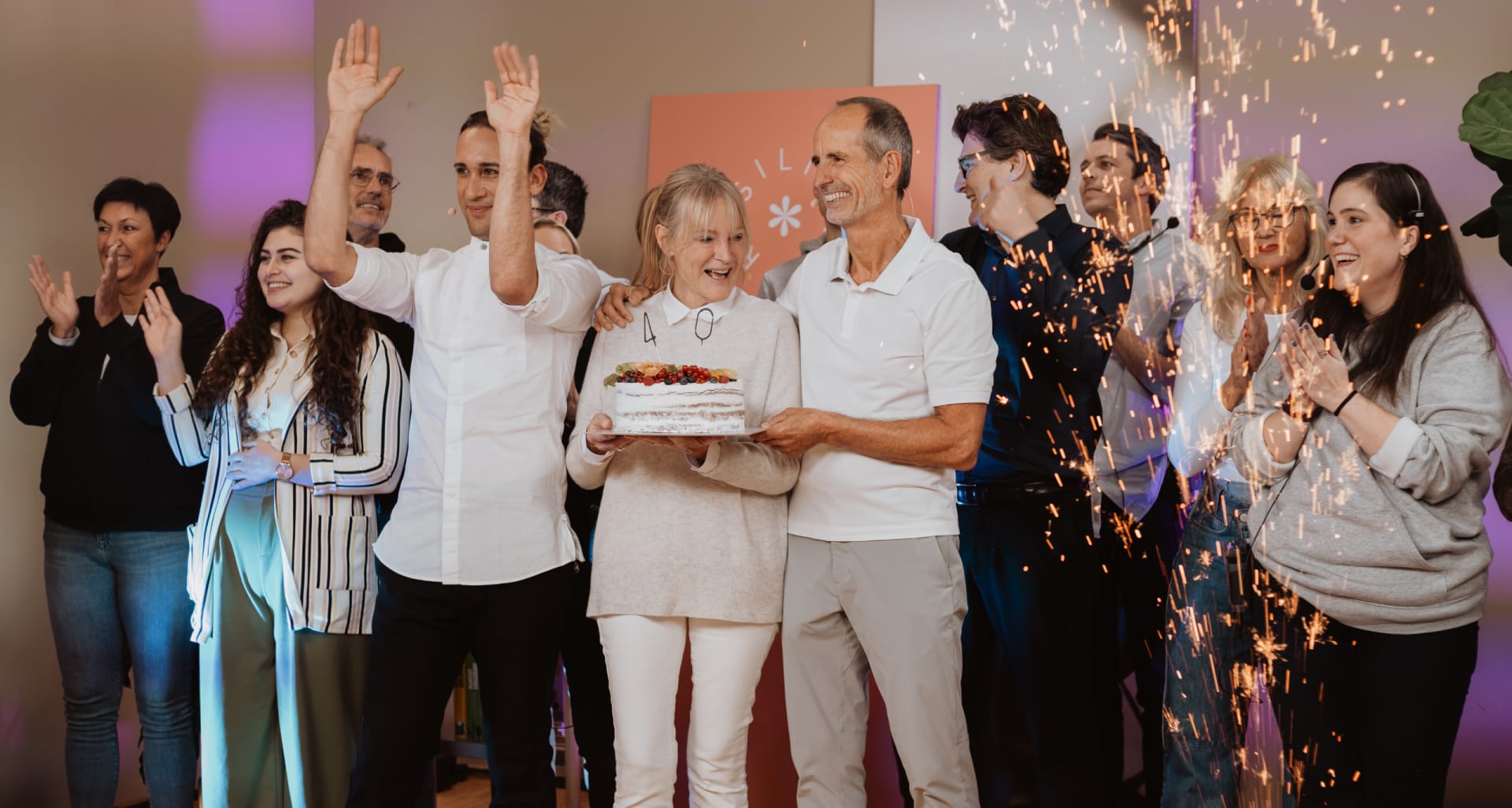 Eine Gruppe von Menschen zelebriert. Im Vordergrund stehen Roland Liebscher-Bracht udn Dr. med. Petra Bracht. Petra hält einen Kuchen in der Hand. Rechts im Bild sieht man eine Funkenfontäne. Die Atmosphäre des Bildes ist glücklich und ausgelassen. Die Menschen im Bild feiern.