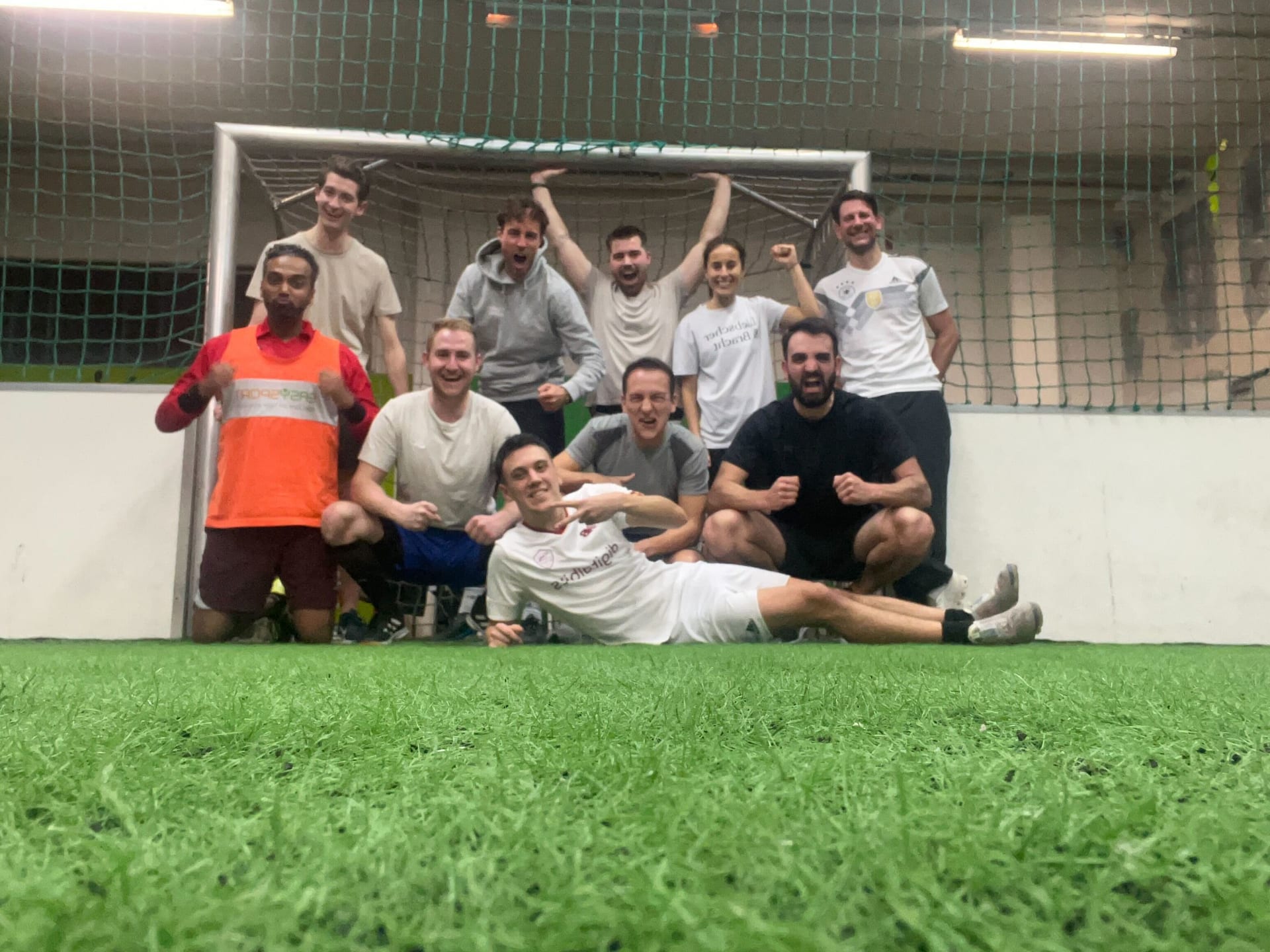 Zu sehen ist eine Gruppe von Menschen vor einem Fußballtor. Sie tragen Sportklamotten und schauen jubelnd in die Kamera. Das Bild vermittelt ein Gefühl von Teamgeist und Sportlichkeit.