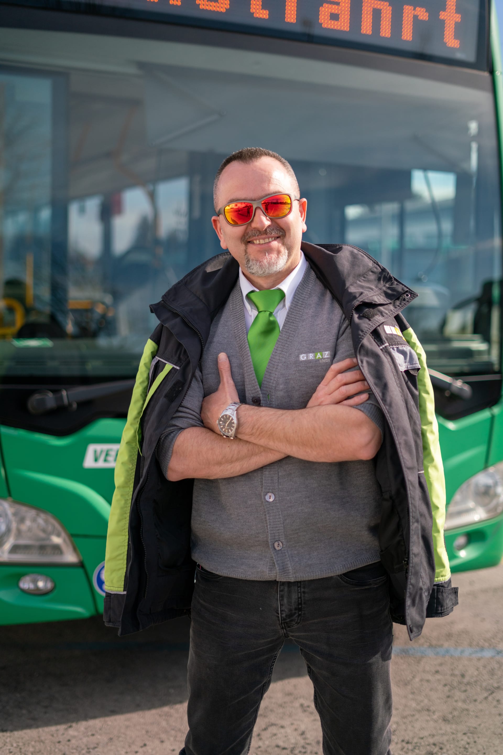 Ein freundlicher Busfahrer der Graz Linien steht mit verschränkten Armen vor einem grünen Bus und trägt eine Sonnenbrille sowie eine graue Uniform mit grüner Krawatte.