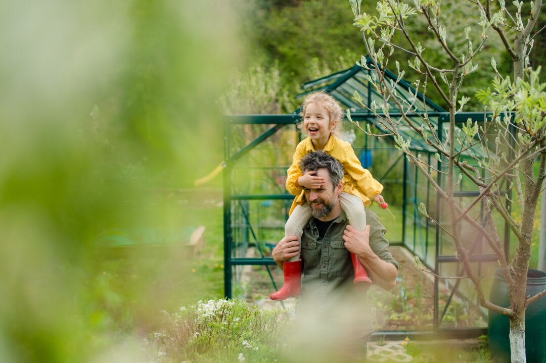 Een klein meisje in regenjas en laarsjes zit bij haar vader op zijn schouders en lacht