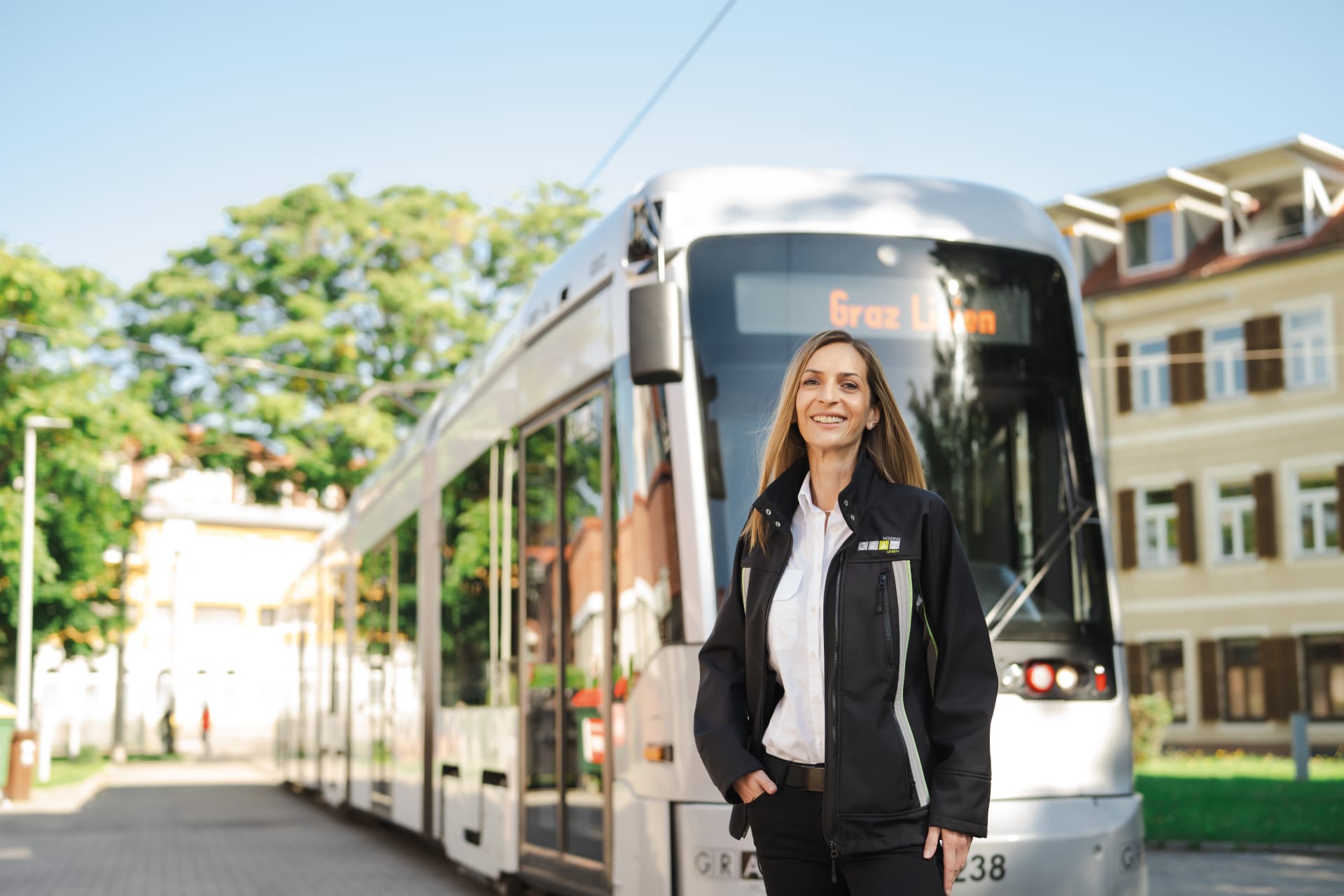 Eine lächelnde Straßenbahnfahrerin der Graz Linien steht vor einer silbernen Straßenbahn in einer sonnigen Umgebung mit Bäumen und Gebäuden im Hintergrund.