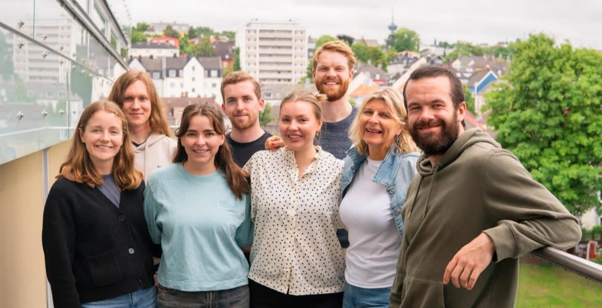 6 smilende studenter og vår daglige leder Marit og fast ansatt Tobias som holder rundt hverandre og smiler utenfor kontoret i Trondheim