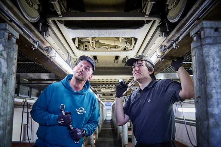 Zwei junge Werkstatt-Mitarbeiter befinden in der Werkstatt-Grube unterhalb einer Straßenbahn. Sie tragen Sicherheitskappen und schauen sich mit einer Taschenlampe den Unterbau der Bahn an.