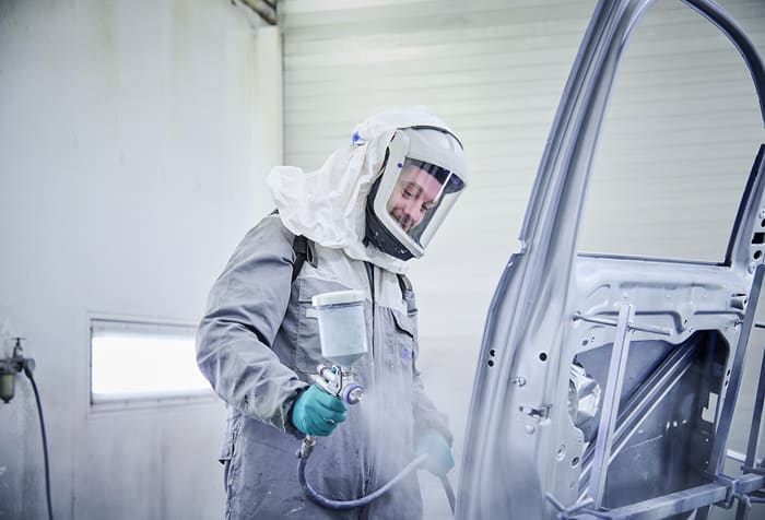 Ein Mitarbeiter steht in der Lackiererei und trägt einen hellen Schutzanzug mit einer Haube. Mit einer Lackierpistole in der Hand lackiert er die geöffnete Tür eines Fahrzeugs.