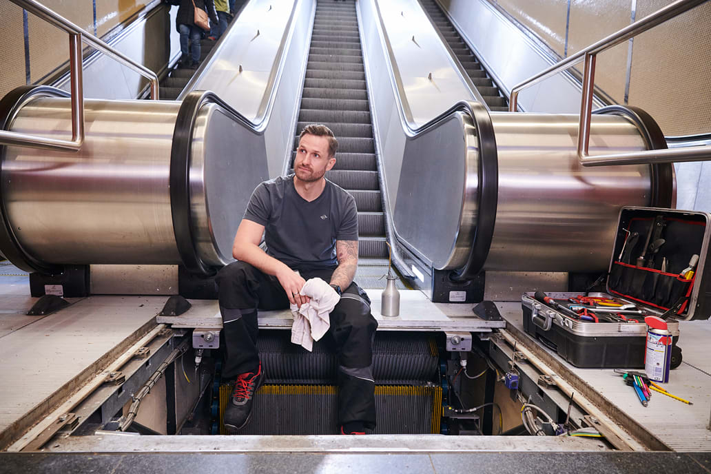 Ein Mitarbeiter der BOGESTRA sitzt in Arbeitskleidung am unteren Ende einer Fahrtreppe. Seine Beine hängen in den geöffneten Schacht vor dem Aufgang der Fahrtreppe. Um ihn herum befinden sich verschiedene Werkzeuge.