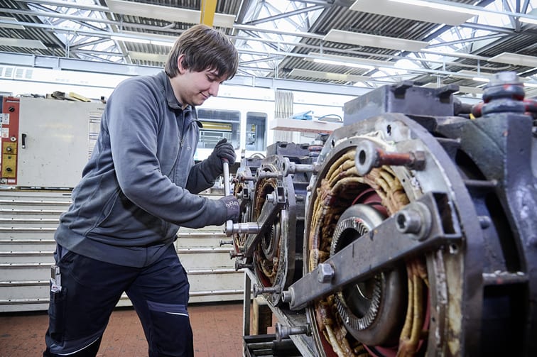 Ein Werkstatt-Mitarbeiter arbeitet mit einer großen Ratsche an dem Fahrwerk einer Straßenbahn.
