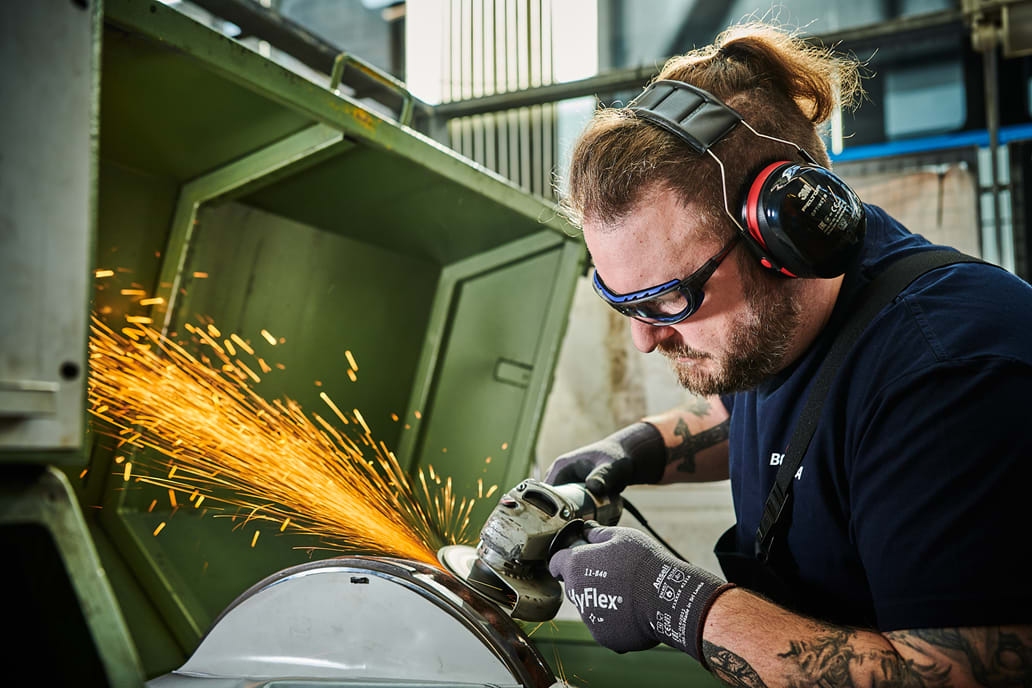 Ein Werkstatt-Mitarbeiter in Sicherheitskleidung setzt eine Flex an ein Werkstück, dabei fliegen viele Funken. Er trägt dabei eine Schutzbrille, Handschuhe und Ohrenschützer.