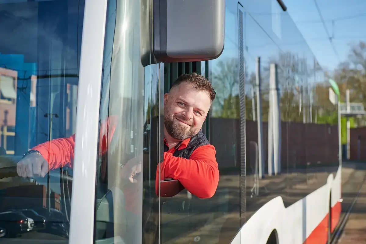 Junger, männlicher Busfahrer in rot-schwarzer Kleidung, der am Steuer eines Busses sitzt, beugt sich aus dem Seitenfenster und lächelt freundlich in die Kamera