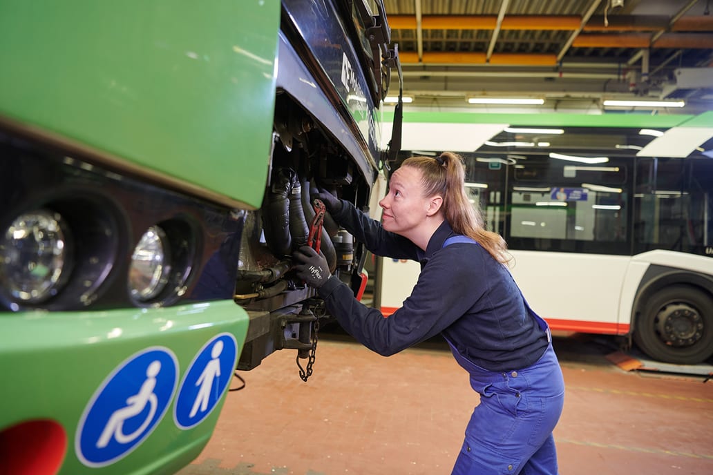 Eine Kfz-Mechatronikerin schaut in den Motorraum eines Busses. Sie hat ein Zange in der Hand.