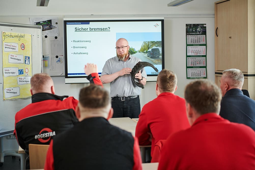 Ein Fahrlehrer steht vor einer Gruppe Fahrschülern und hält das Profil eines Reifens in der Hand. Ein Fahrschüler meldet sich.