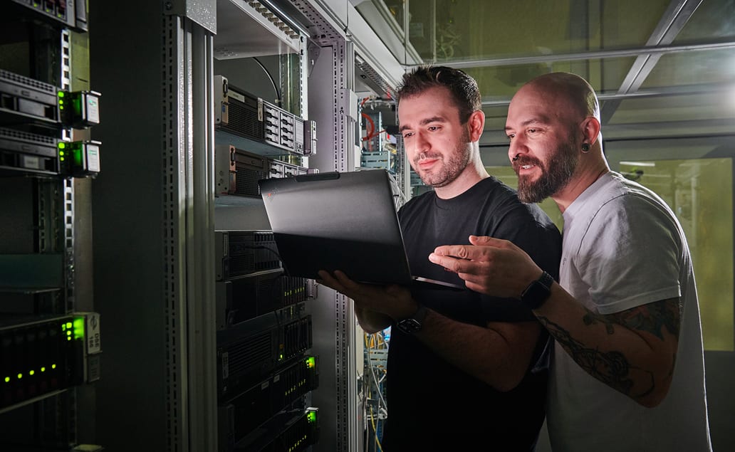 Zwei junge IT-Mitarbeiter der BOGESTRA stehen im Serverraum neben einer Reihe an Servertürmen. Einer hält einen Laptop in der Hand, auf den beide gemeinsam schauen.