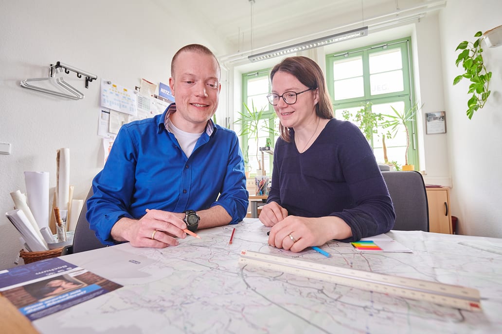 Ein Mann und eine Frau sitzen in einem hellen Büro an einem Tisch, beugen sich über eine große Karte und halten dabei Stifte.