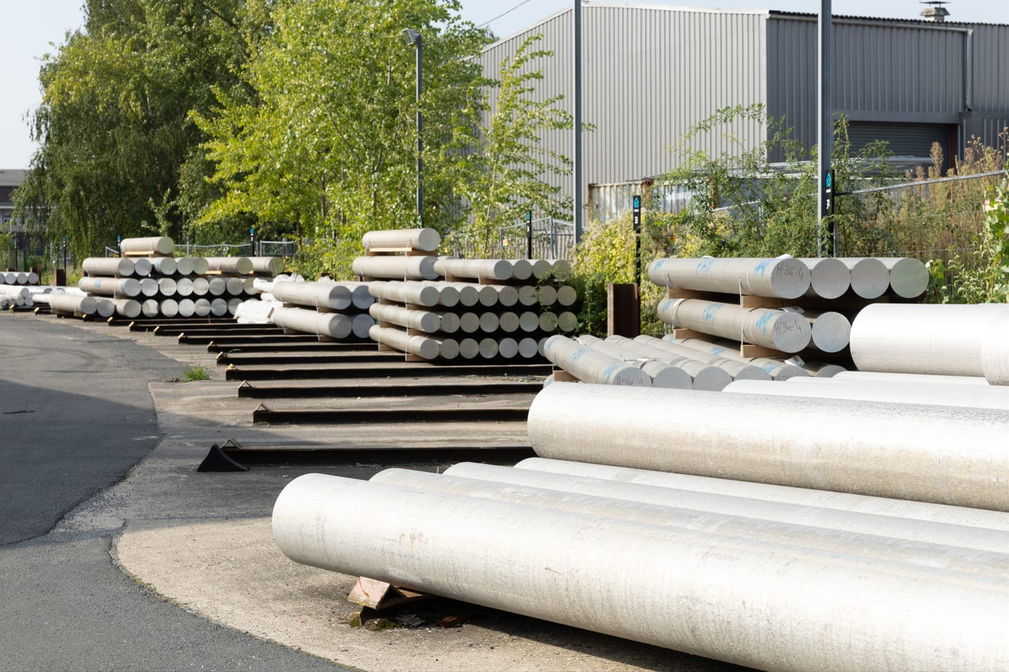 Mehrere Aluminiumbarren sind ordentlich in Stapeln gelagert. Die Barren dienen als Vormaterial für die weitere Verarbeitung und stammen von externen Gießereien. Sie werden zur Herstellung von Aluminiumprodukten eingeschmolzen und bilden den Ausgangsstoff für nachfolgende Produktionsschritte.