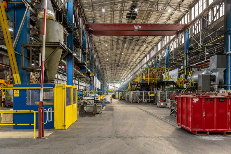 Die Aufnahme zeigt die Produktionshalle. In der Halle sind verschiedene Maschinen, Anlagen und Arbeitsbereiche für die Herstellung und Bearbeitung von Aluminiumprofilen angeordnet.