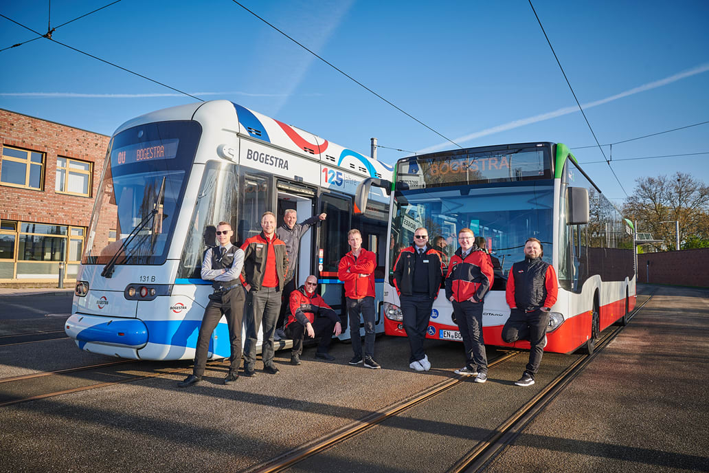 7 Männer in Dienstkleidung stehen vor einer Straßenbahn und einem Bus, ein weiterer Fahrer sitzt in der Tür der Straßenbahn zwischen den anderen Männern.