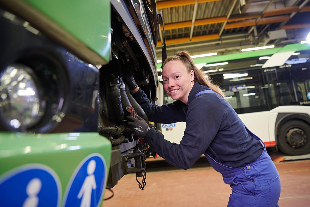 Eine Werkstattmitarbeiterin steht gebeugt vor dem geöffneten Motorraum eines Busses. Sie hält eine Zange in der Hand.