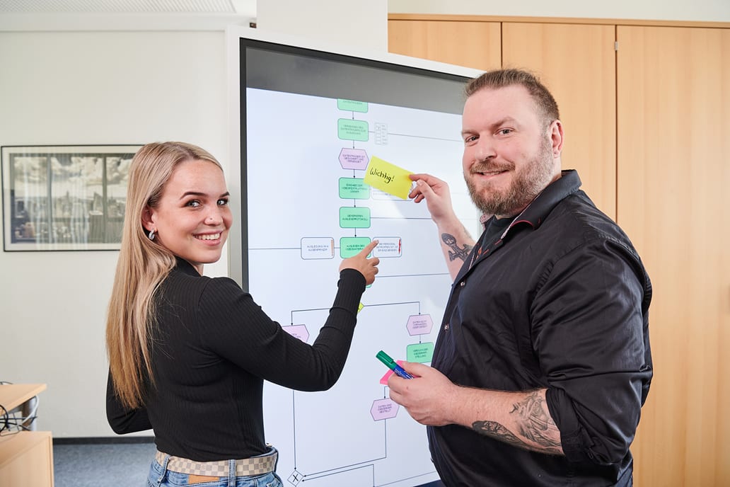 Eine junge Mitarbeiterin und ein Mitarbeiter stehen vor einem großen Bildschirm. Er hlt eine Post-it in der einen und einen Stift in der anderen Hand. Sie zeigt mit dem Finger auf das Post-it.