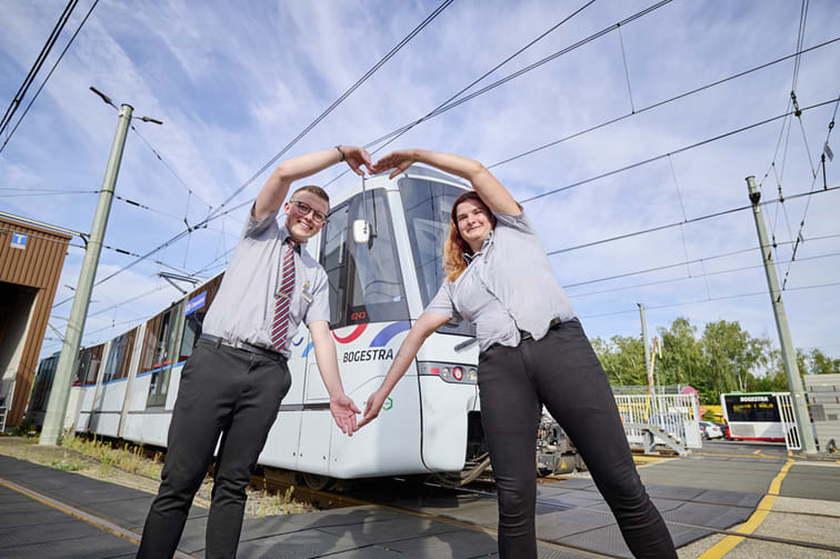 Zwei junge Mitarbeitende in Dienstkleidung stehen vor einer Straßenbahnen und formen mit ihren Armen ein Herz.