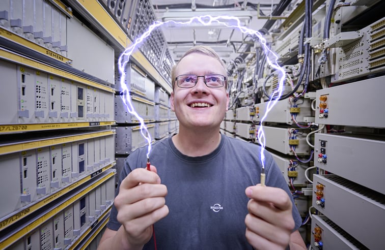 Ein junger Mann steht zwischen 2 langen Reihen mit Steuerungskästen. In den Händen hält er jeweils ein Kabel. Zwischen den Kabeln wurde künstlich ein Lichtbogen erzeugt.