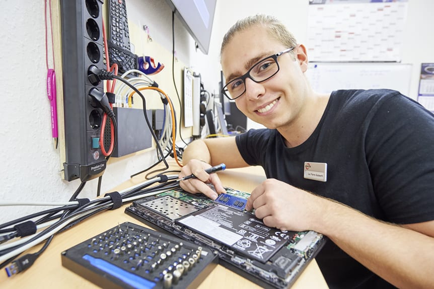 Ein junger IT-Mitarbeiter sitzt vor einem aufgeschraubten Laptop. Er hält einen kleinen Schraubendreher in der Hand.