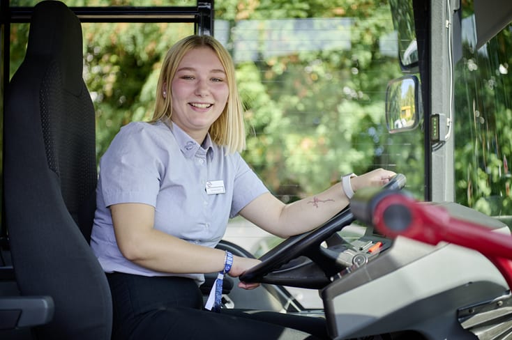 Junge Frau sitzt am Steuer eines Busses und schaut zur Seite in Richtung der vorderen Einstiegstür. Sie hält das Lenkrad mit beiden Händen fest.