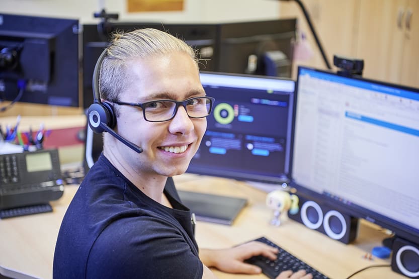 Ein junger Mann sitzt an einem Schreibtisch und hat die Hände auf einer Computertastatur. Er hat ein Headset auf. Im Hintergrund sind zwei Bildschirme und ein Telefon zu sehen.
