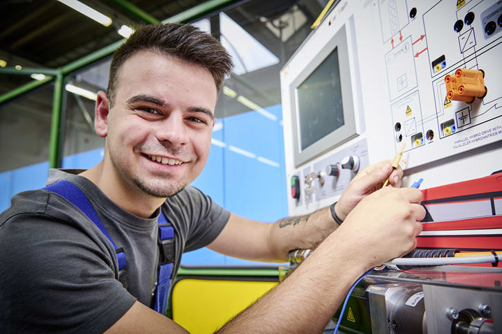 Ein junger Mann in Blaumann steht vor einer Hochvolt-Schulungswand. Er hält zwei Kabel in der Hand.