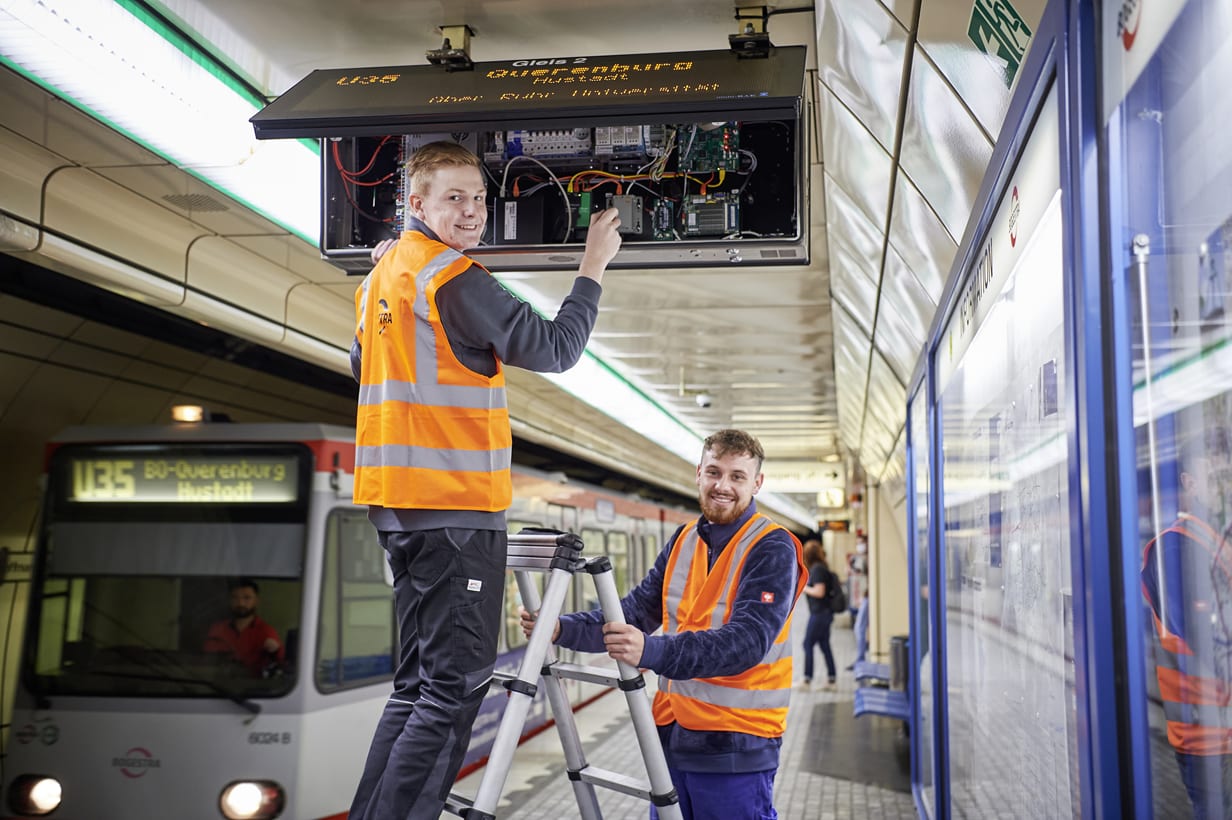 Zwei junge Männer in Sicherheitsweste sind in einem U-Bahnhof. Einer steht auf einer Leiter und arbeitet an einem aufgeklappten Abfahrtsmonitor. Der zweite hält die Leiter fest.
