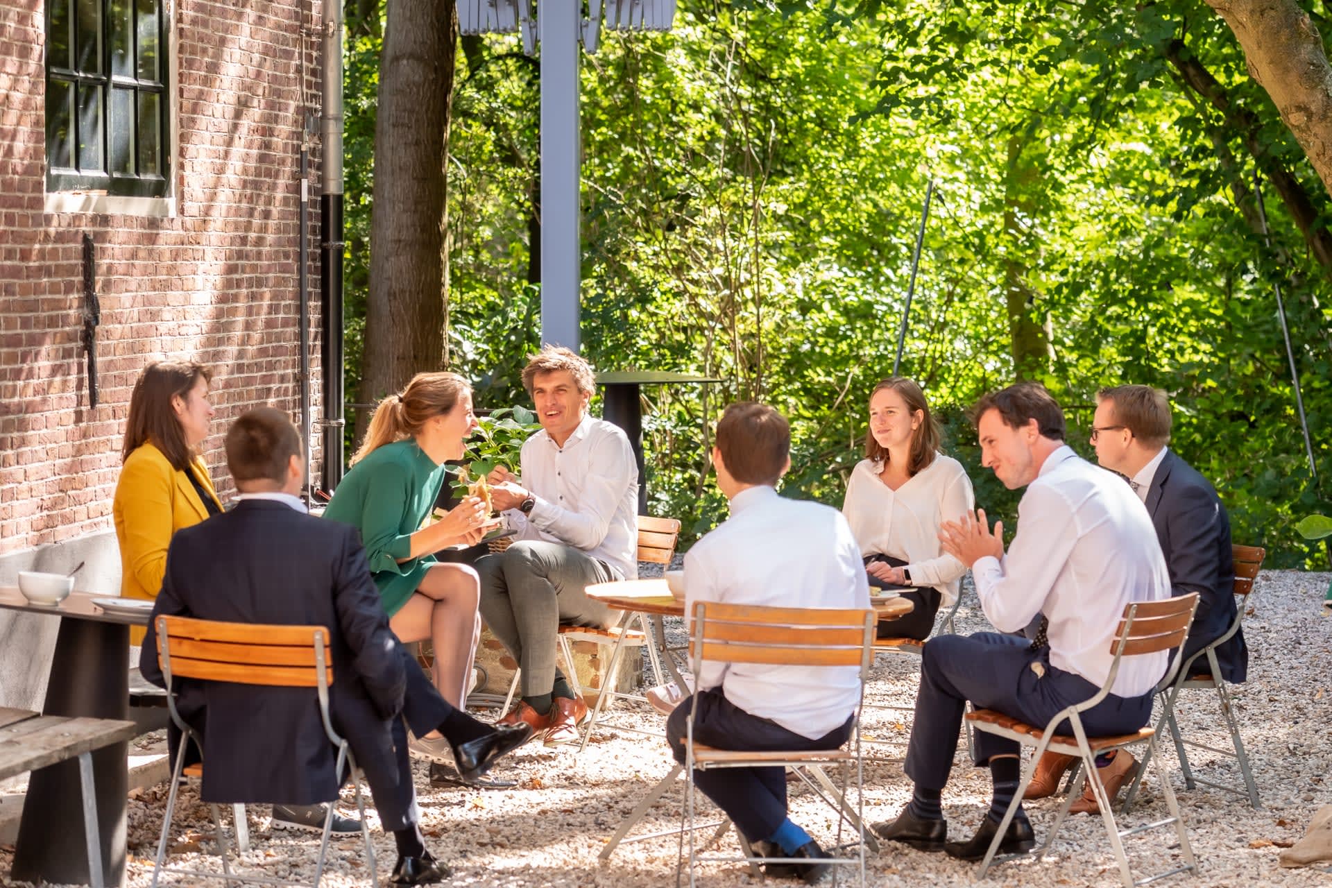 Eight consultants having lunch outside in the garden, talking and laughing with each other. Sitting on chairs in green surroundings.