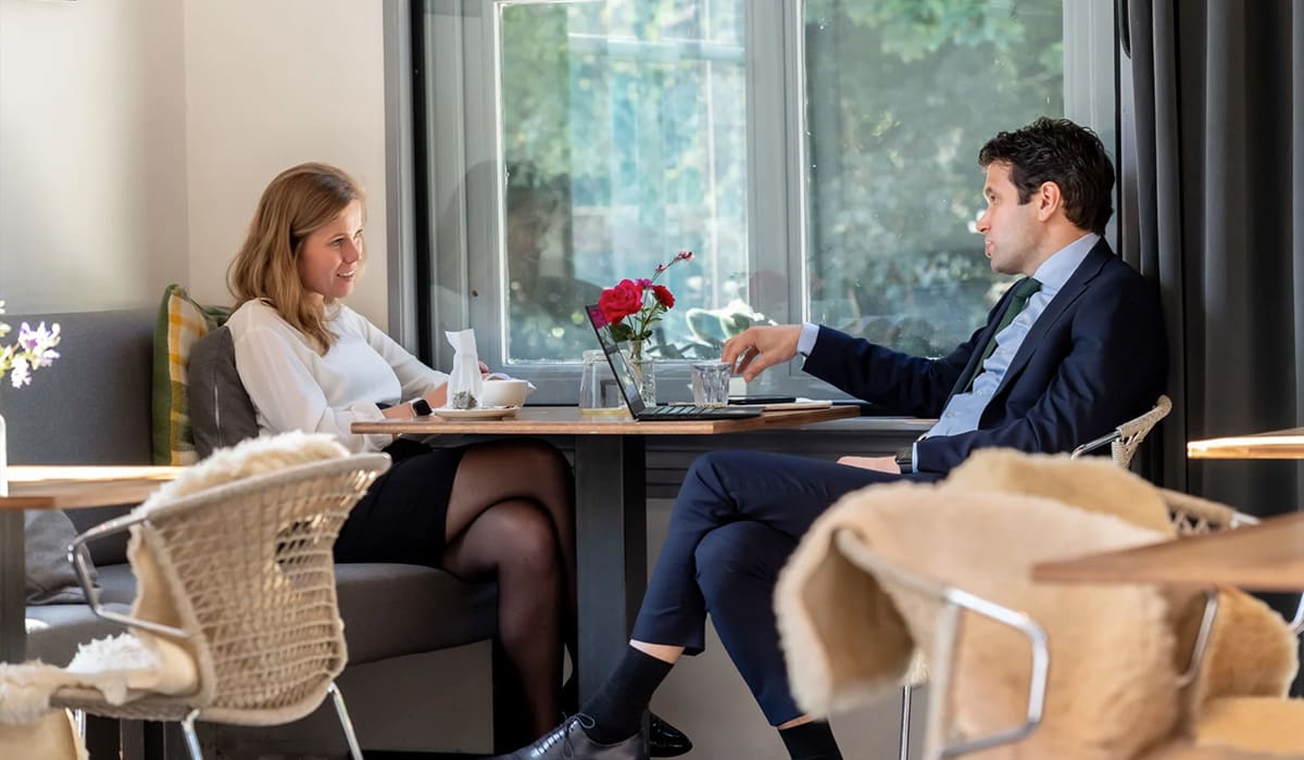 Twee collega's die met elkaar praten aan een tafel, met koffie, water en laptop. 
